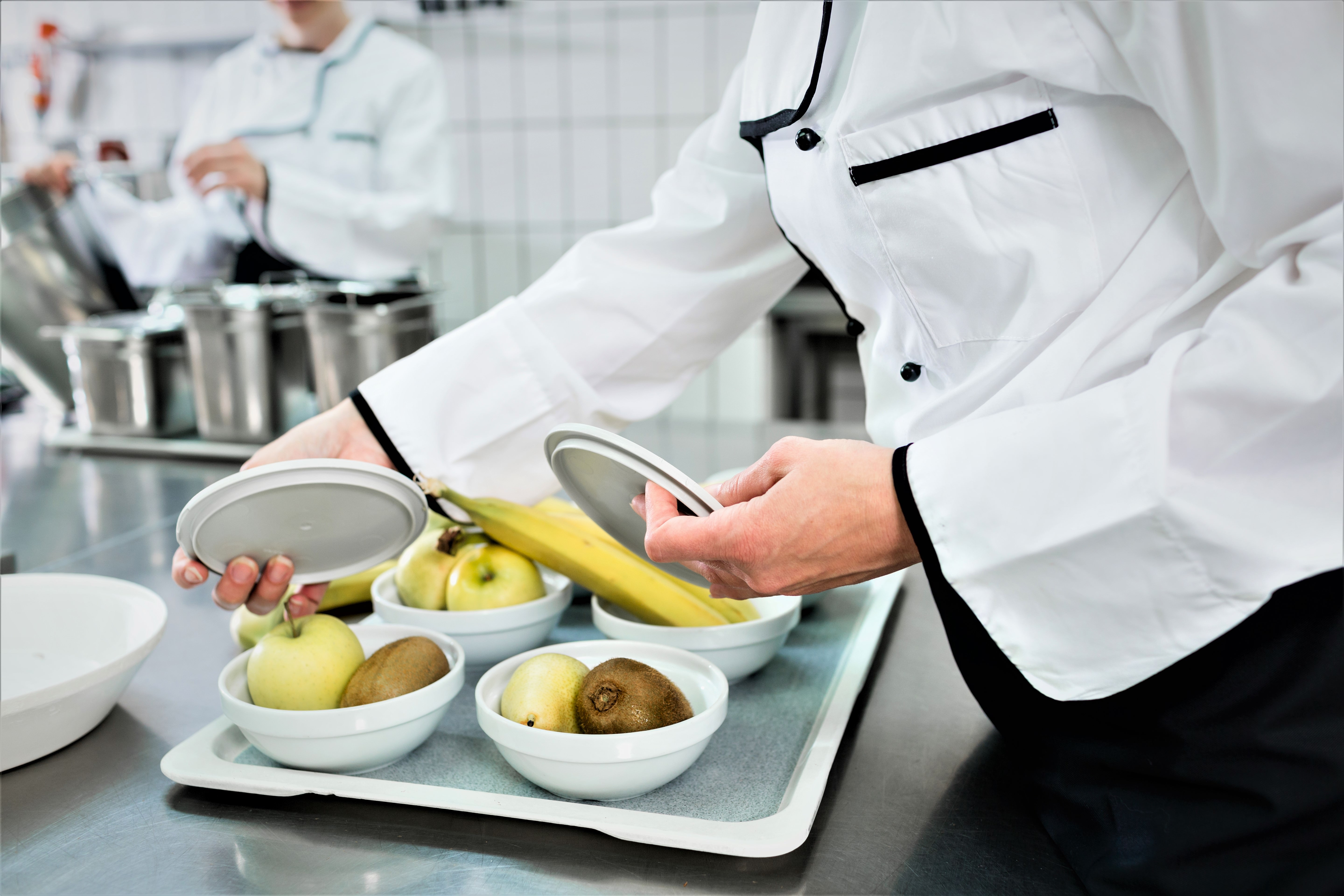 En kok er ved at anrette skåle med frugt; bananer, kiwier og æbler, i et køkken.