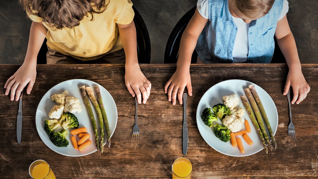 To børn sidder ved et spisebord med tallerkener fyldt med grønsager