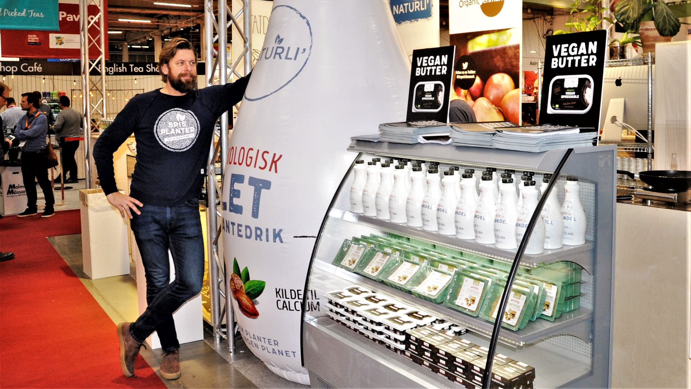 Henrik Lund fra Naturli' Foods står på en fødevaremesse op ad en oppustelig flaske. Ved siden af er en køler med nogle af virksomhedens produkter.