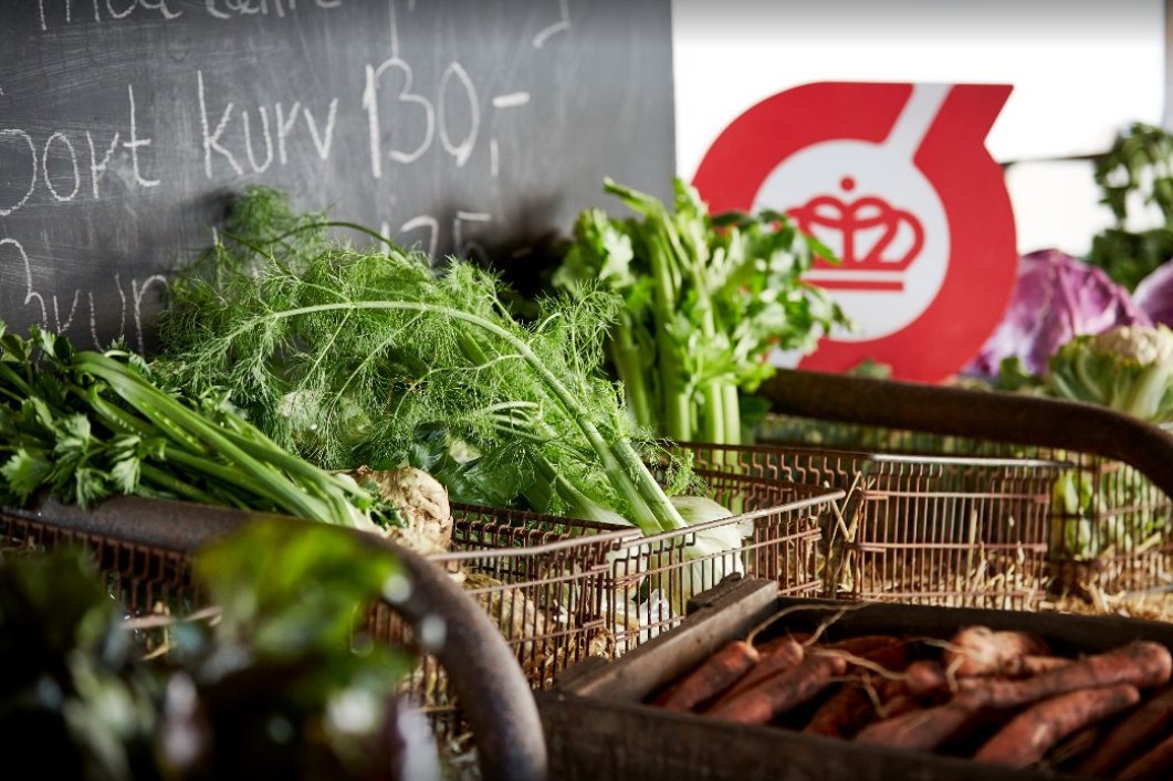 Bakker med frisk frugt og grønt står foran en pristavle. I baggrunden ses det røde Ø-mærke.