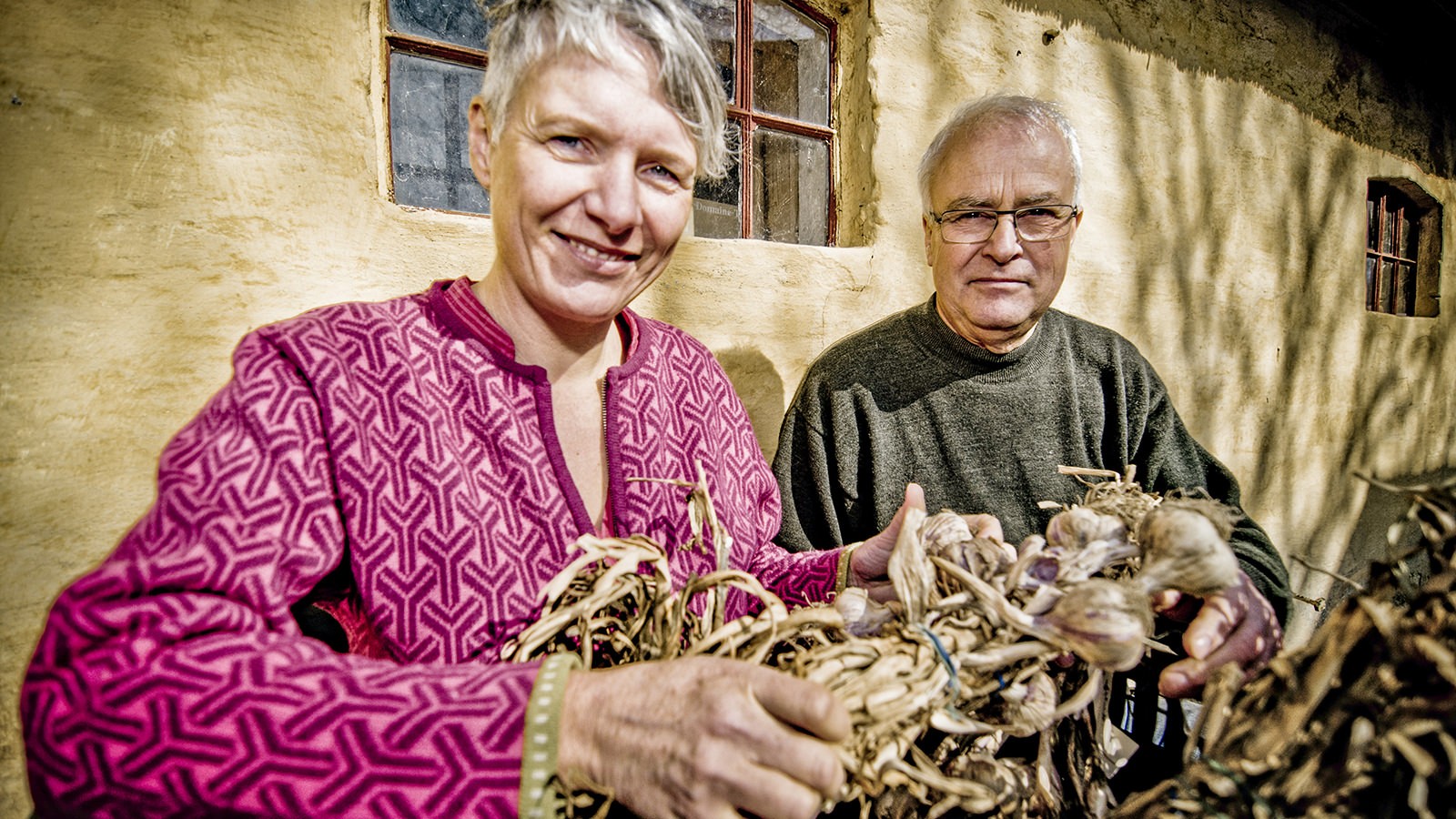 Lotte og Peter Lei står foran deres gård med et bundt hvidløg i hænderne.