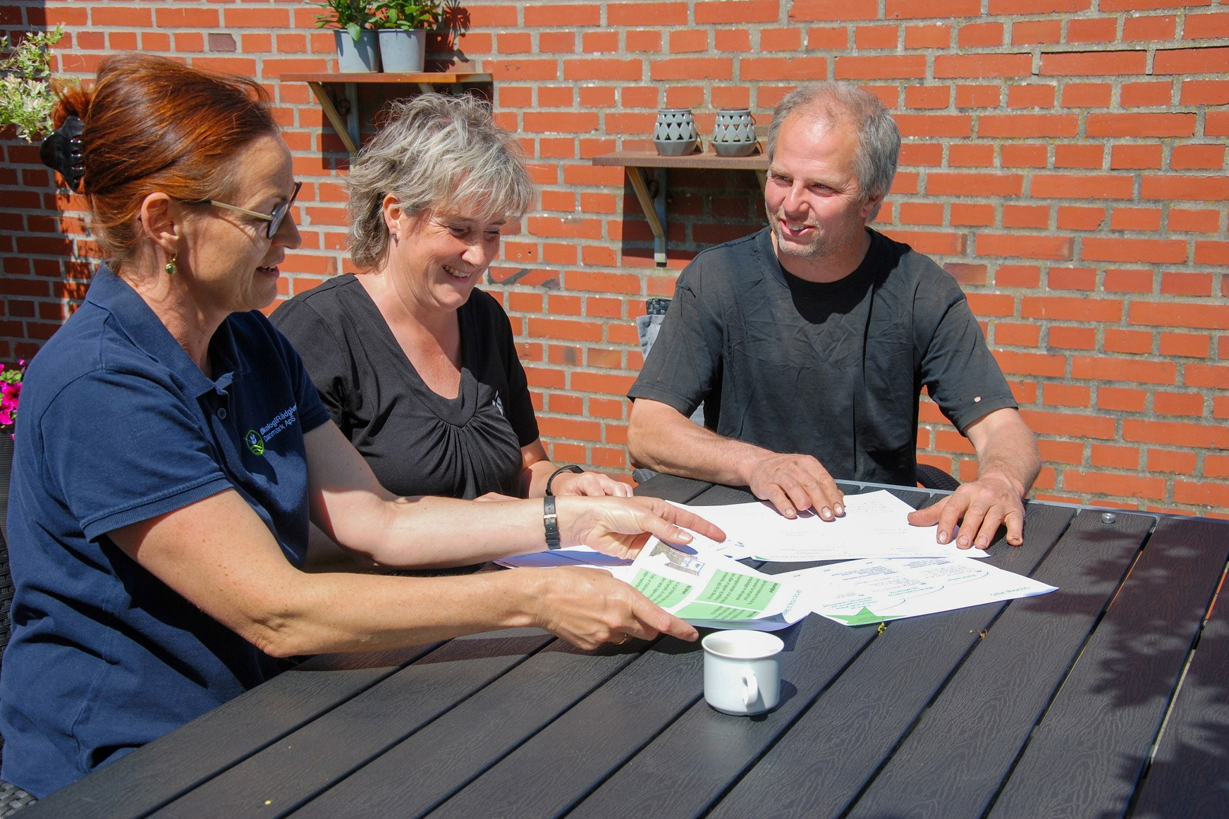 Solvejg Horst sidder ved et havebord og viser Tina og Jan Bolding nogle papirer