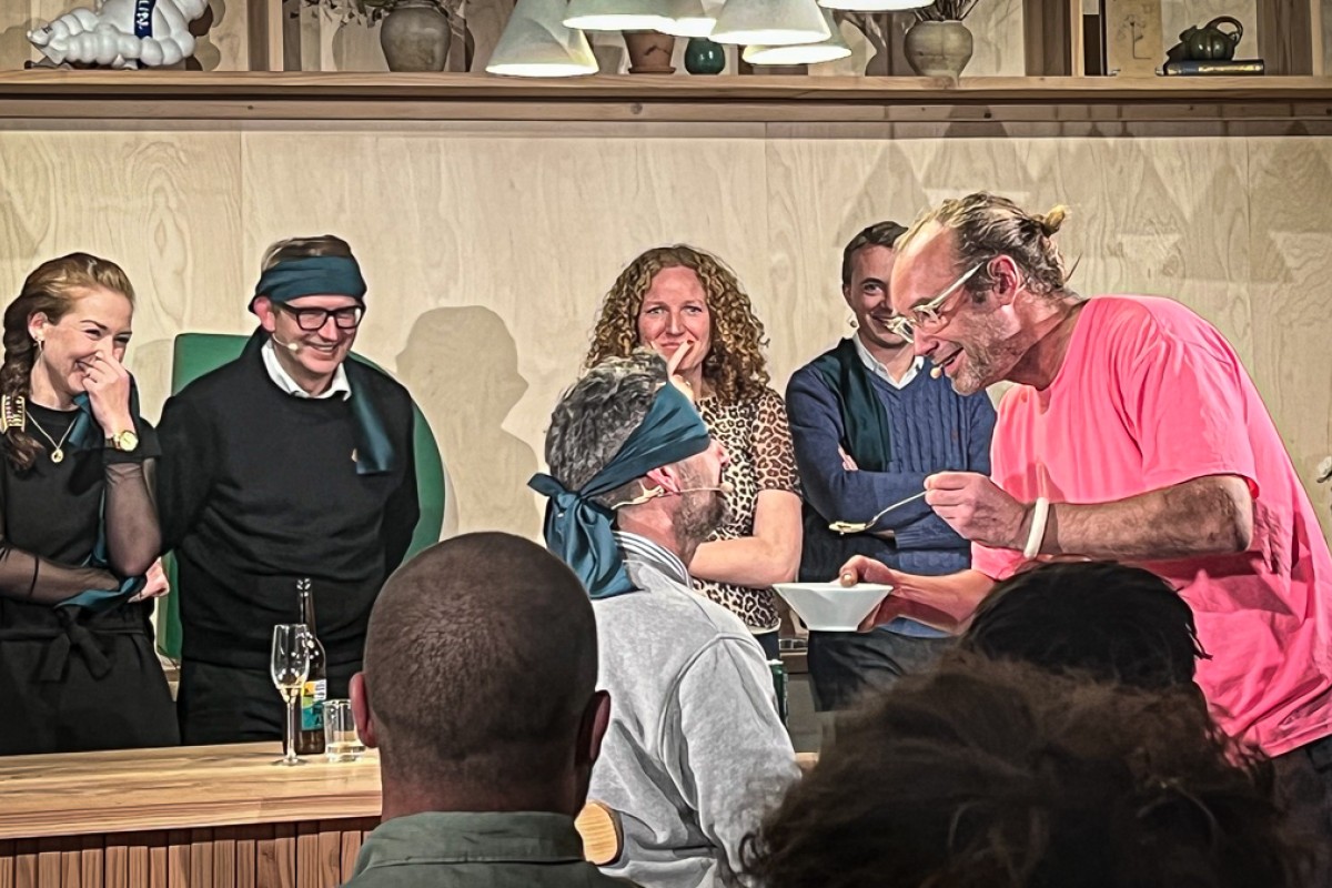 En person med bind for øjnene skal smage på en skefuld mad, mens fire politikere står bagved og smiler