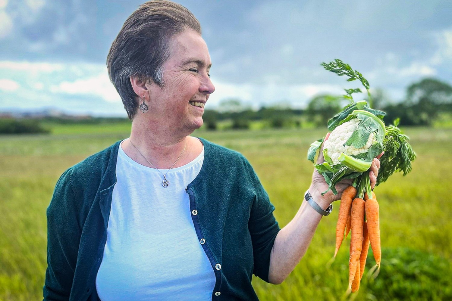Hanne Lakkenborg Kristensen står på en mark med et blomkål og et bundt gulerødder i hånden